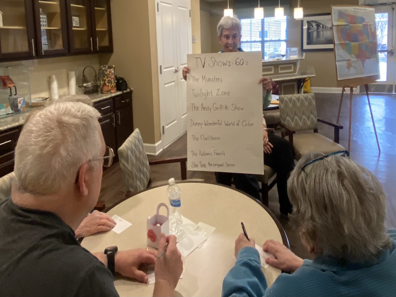 An NEUMC volunteer holds up a board of TV Shows from the 60s to Harmony Assisted Living residents. They appear to be playing a trivia game of sorts.