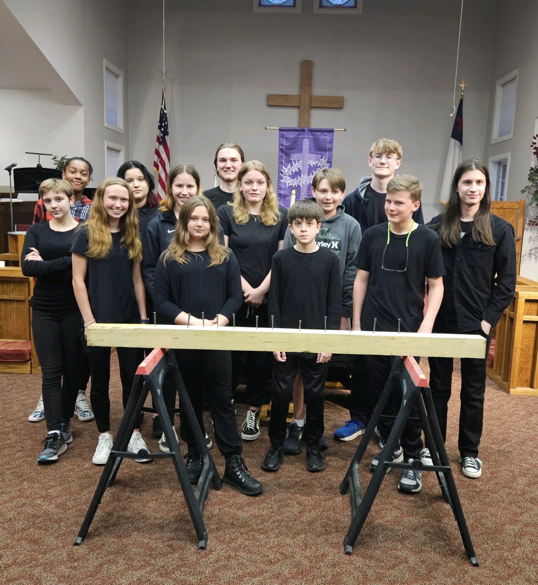 A group of youth members dressed in black stands for a picture after the Good Friday service. In front of them, there is a wooden board propped up on sawhorses with a bunch of nails in it.