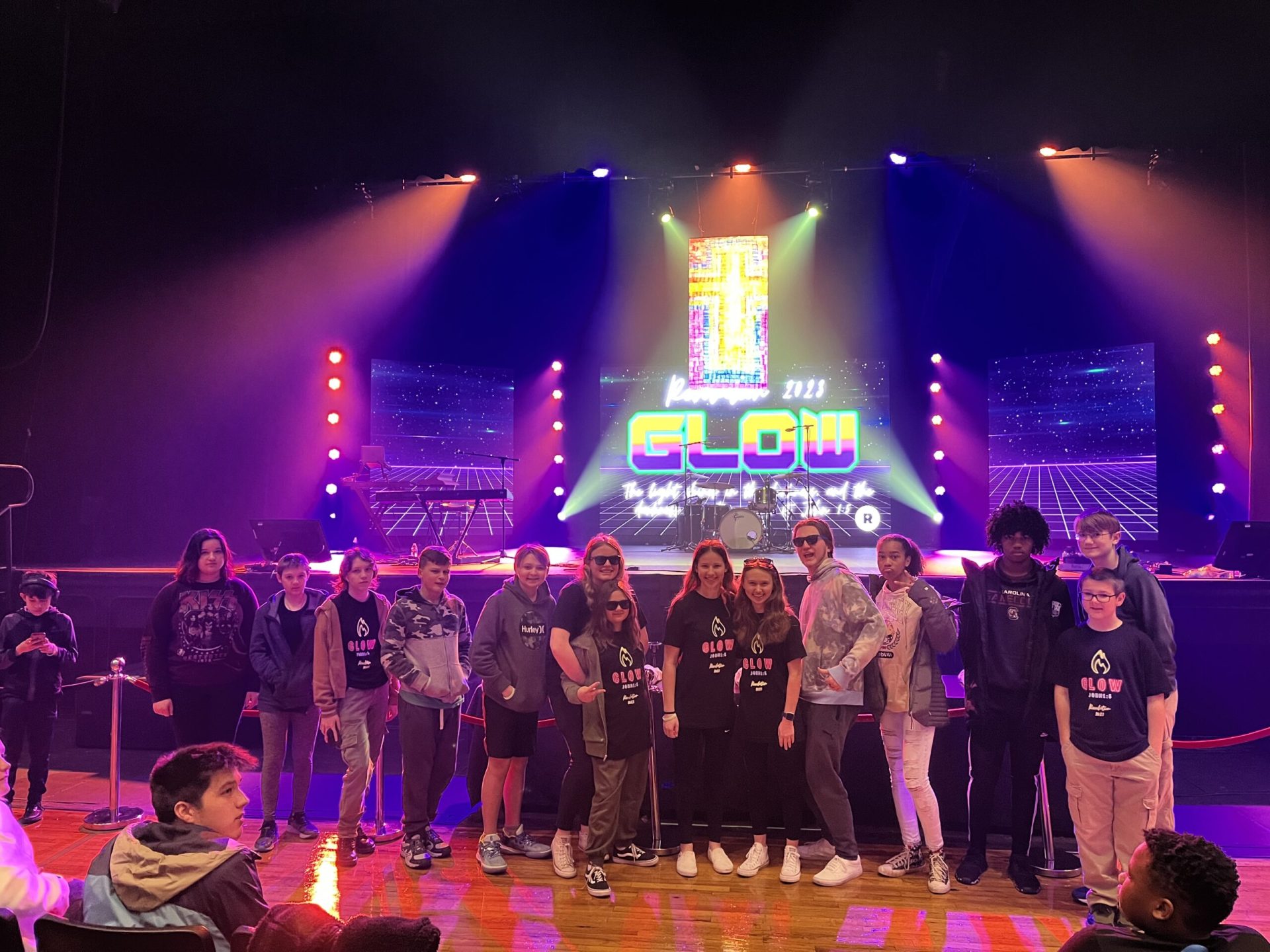 A group of NEUMC youth members, with Revolution 2024 shirts on, pose for a picture in front of the stage at Township Auditorium