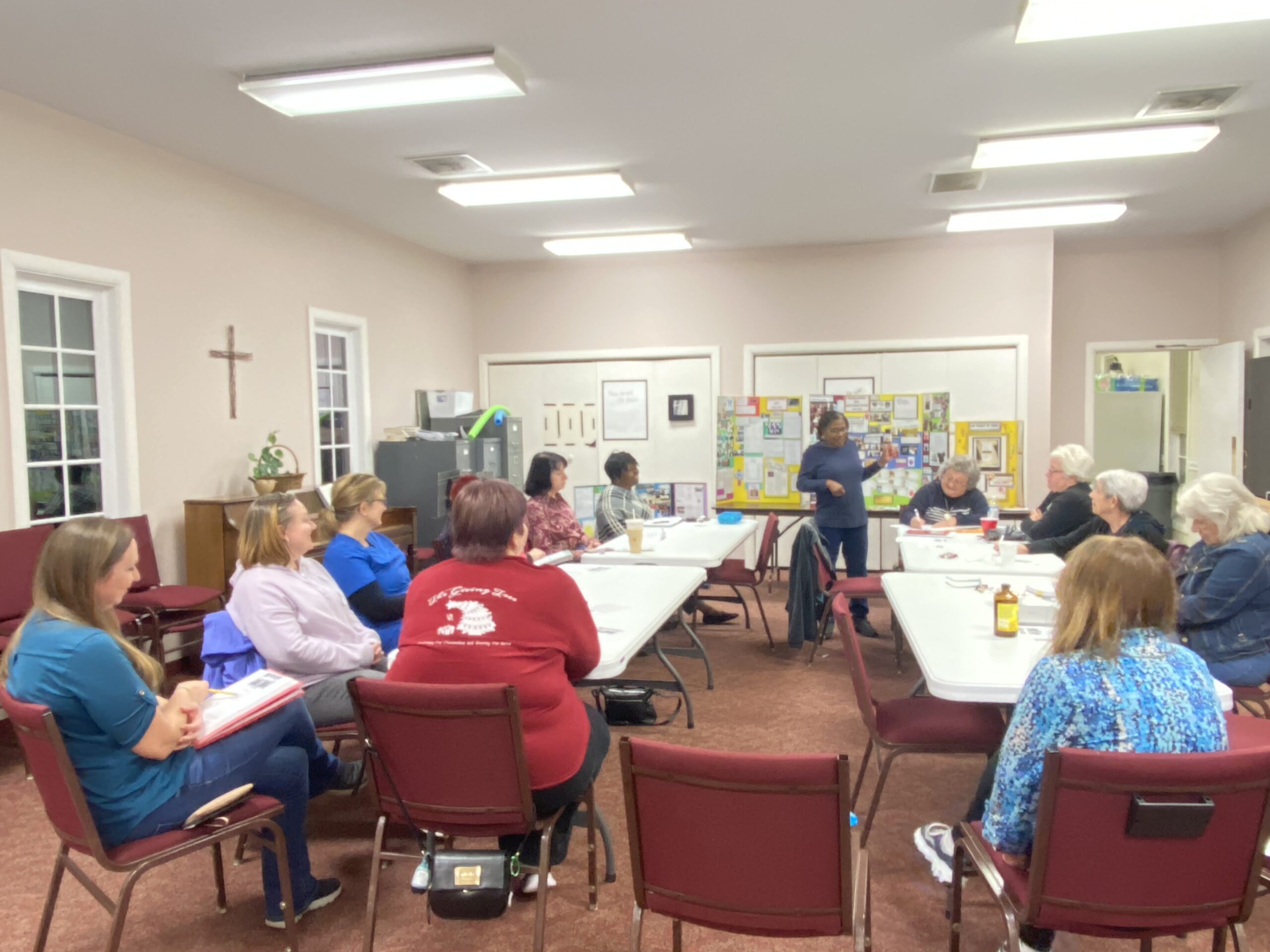 The United Women in Faith Group gathers in Room 102 for an evening meeting. Sylvia stands in front of the group with yellow posterboards presenting information.